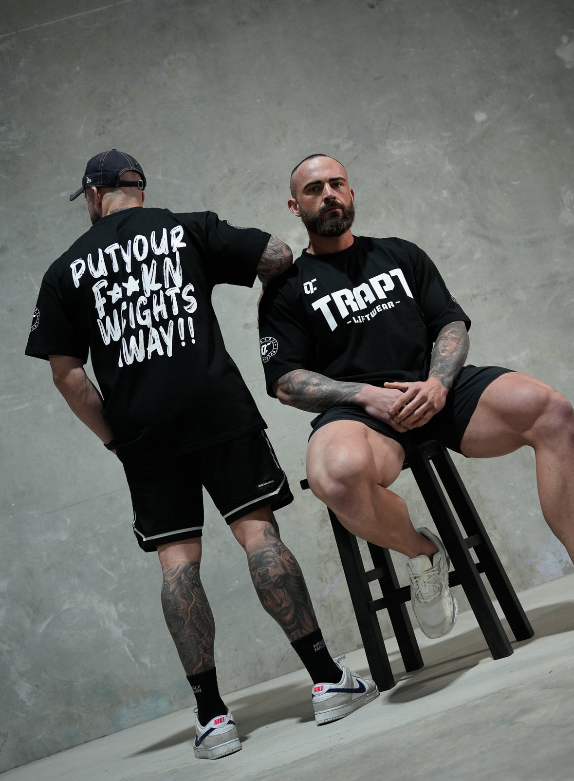 Two men wearing black t-shirts with text, sitting on a stool against a gray concrete wall.