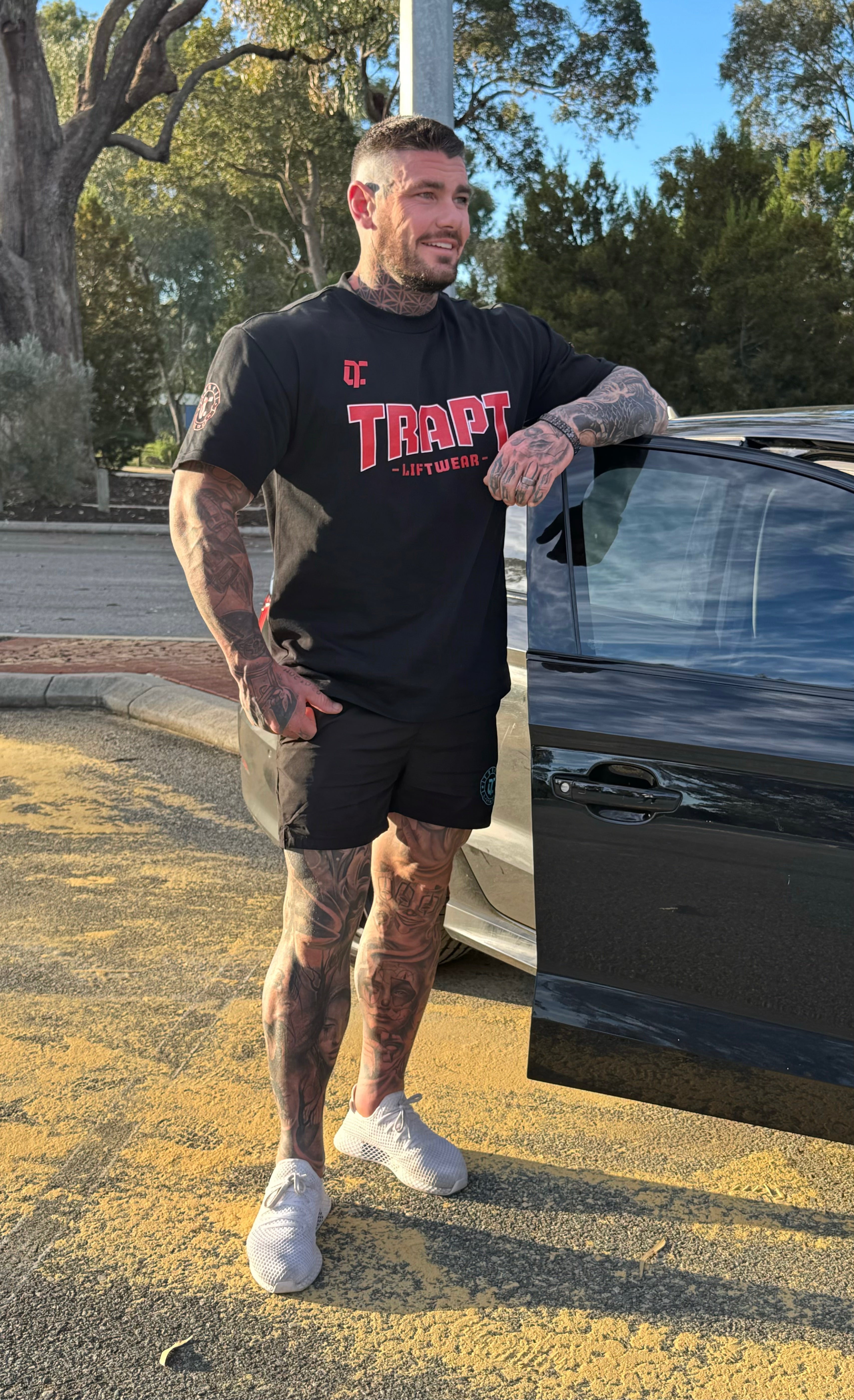 Man with tattoos standing next to a car wearing a black t-shirt with red text.